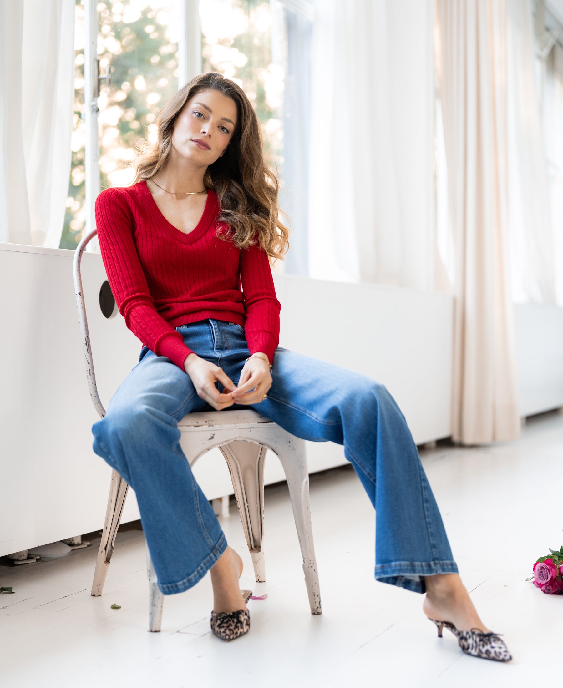 Woman wearing red LA JULYA cable knit V-neck sweater, seated on chair in bright room, looking at camera.