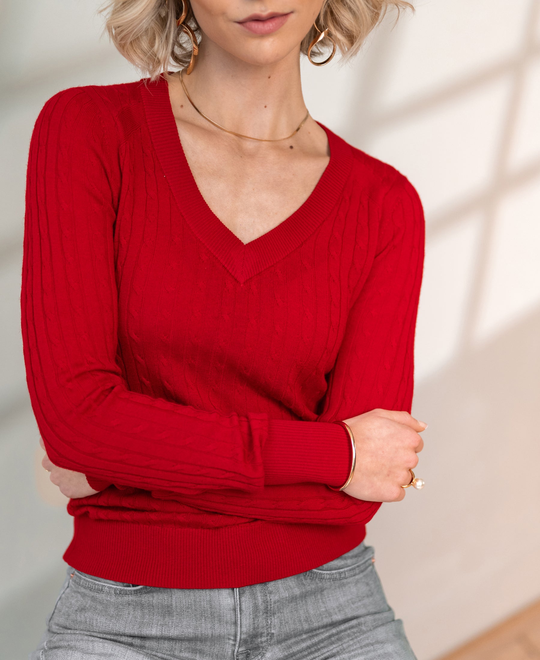 Woman wearing a red LA JULYA cable knit V-neck sweater from OVERIG, arms crossed.