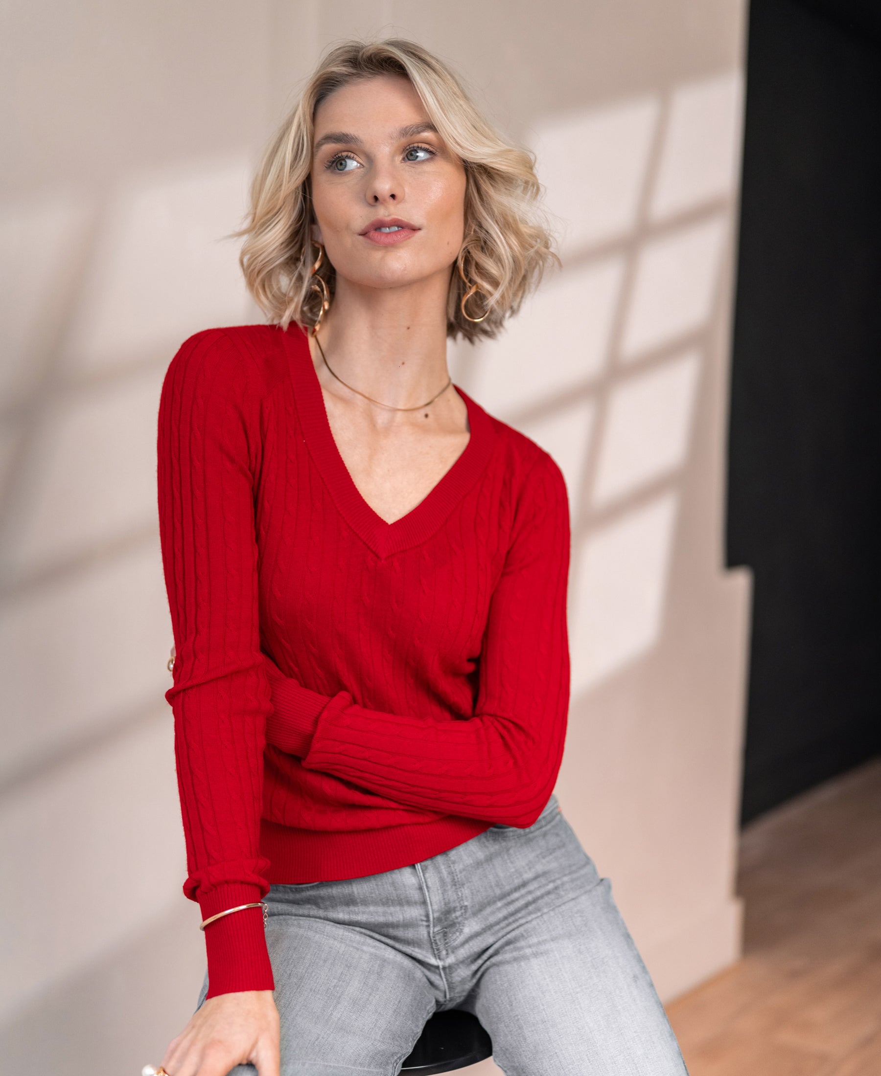 Red LA JULYA V-neck cable knit sweater shown on a woman, with a sunlit wall in the background.