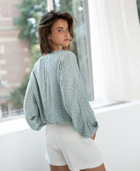 Mist green-ivory polka dot blouse in lightweight fabric, shown from the back as worn by a woman standing by a window.