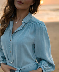 Woman wearing a bleached Tencel shirt dress, standing on the beach at sunset with waves in the background.