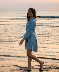 Woman wearing light blue Tencel shirt dress walks along shoreline at sunset, waves in the background.