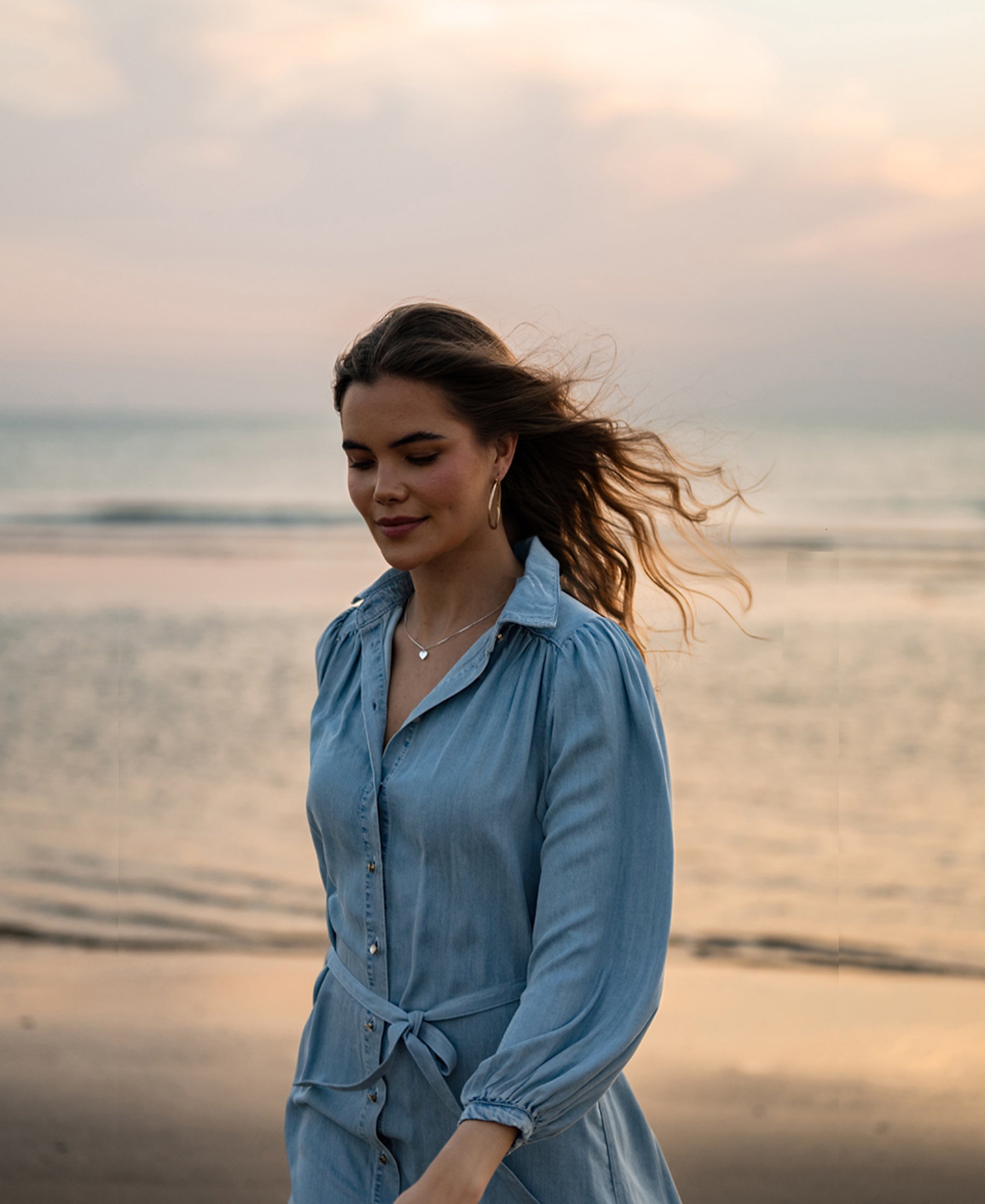 Woman wearing a bleached Tencel shirt dress by PLEIN PUBLIQUE, walking on the beach at sunset with eyes closed.