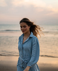 Woman wearing a bleached Tencel shirt dress by PLEIN PUBLIQUE, walking on the beach at sunset with eyes closed.