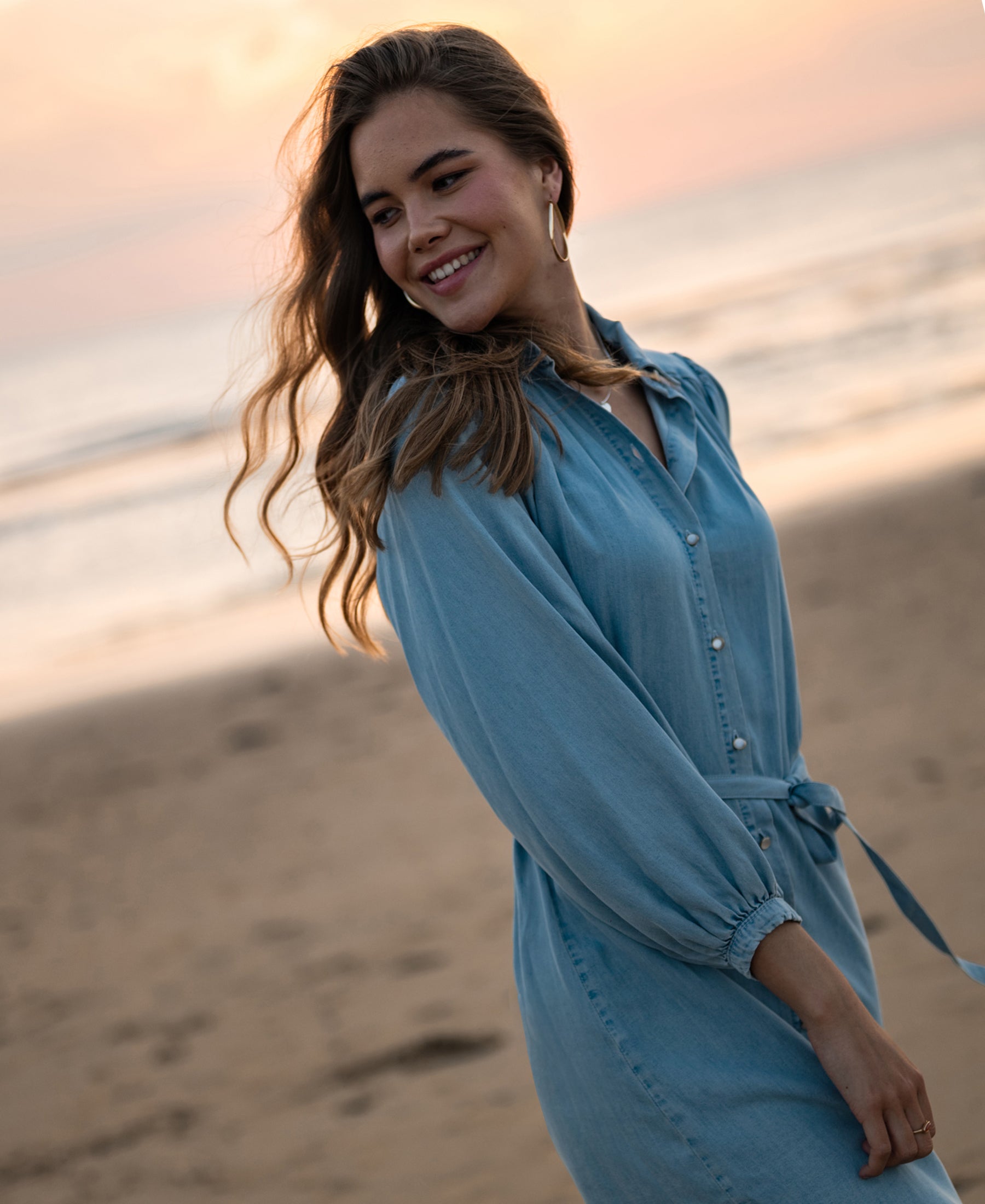 Tencel shirt dress in bleached light color, shown on a sandy beach at sunset with waves in the background.