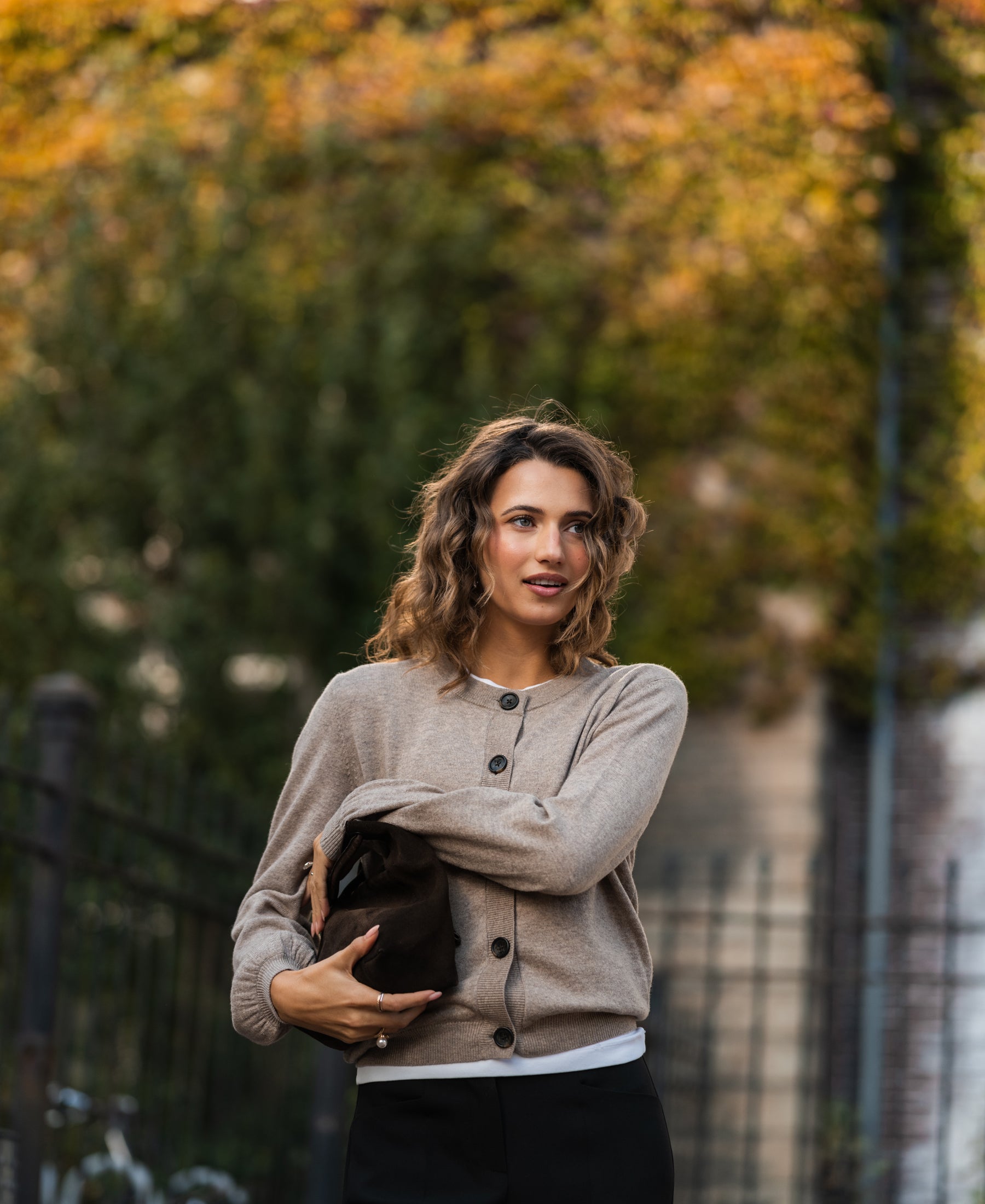 Warm taupe round neck vest sweater by PLEIN PUBLIQUE, shown outdoors with autumn trees in the background.