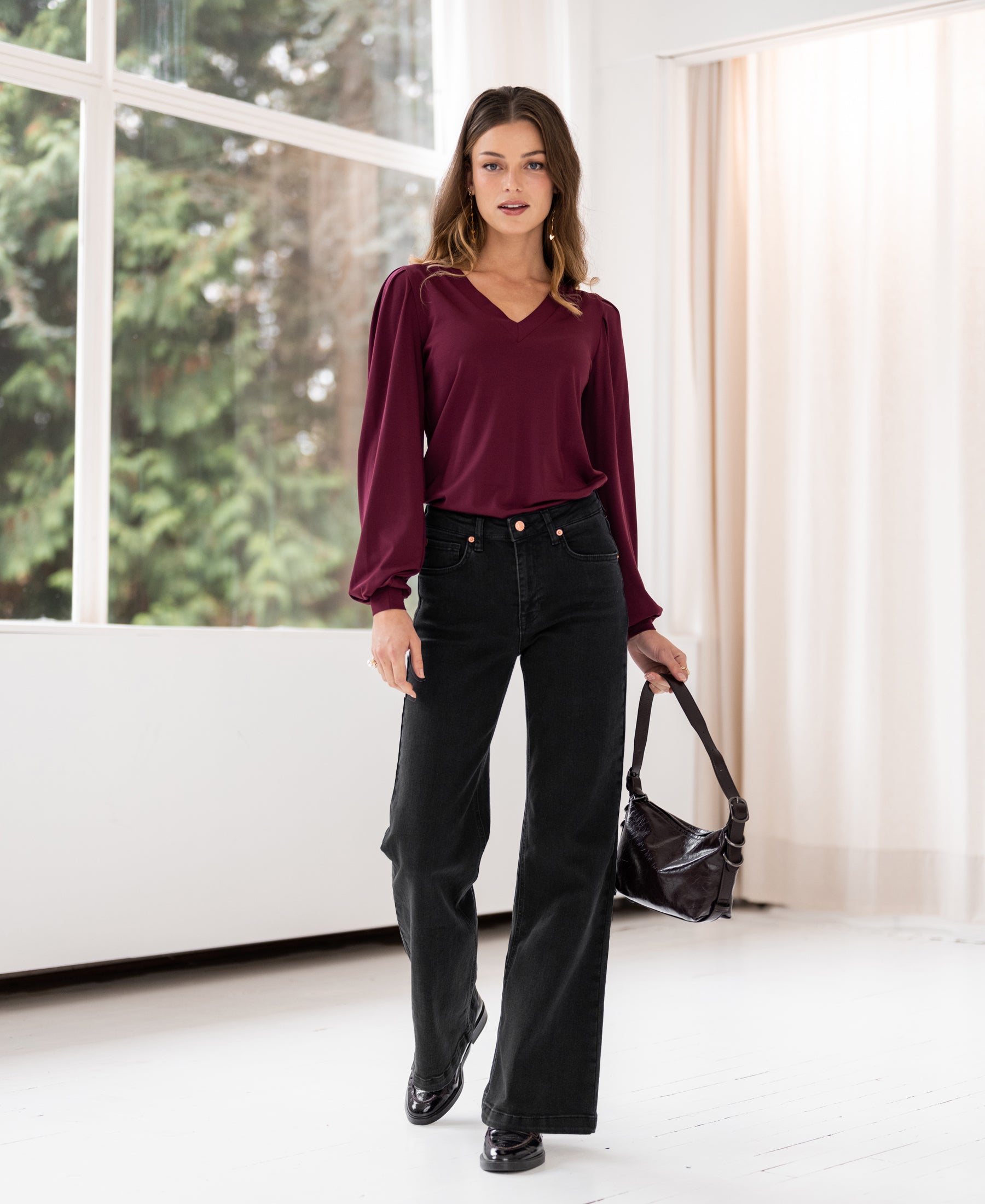 Woman wears the LA LIANA Bordeaux V-neck top by PLEIN PUBLIQUE indoors near large windows.