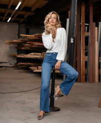 Wide leg stonewash jeans in denim, shown worn by a woman sitting on a stool in a wood workshop.