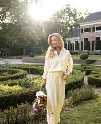 Light yellow LA LYNETTE trousers from OVERIG, shown in a sunlit garden.