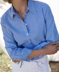 Person wearing the PLEIN PUBLIQUE Striped Blouse LA LOIRE White-Blue, arms crossed outdoors.