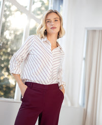 Woman wearing an ivory and wine red striped blouse stands by a window.