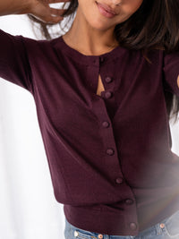 Woman in a PLEIN PUBLIQUE merino wool and silk cardigan, style LA LORAINE, color wine, arm raised against white background.