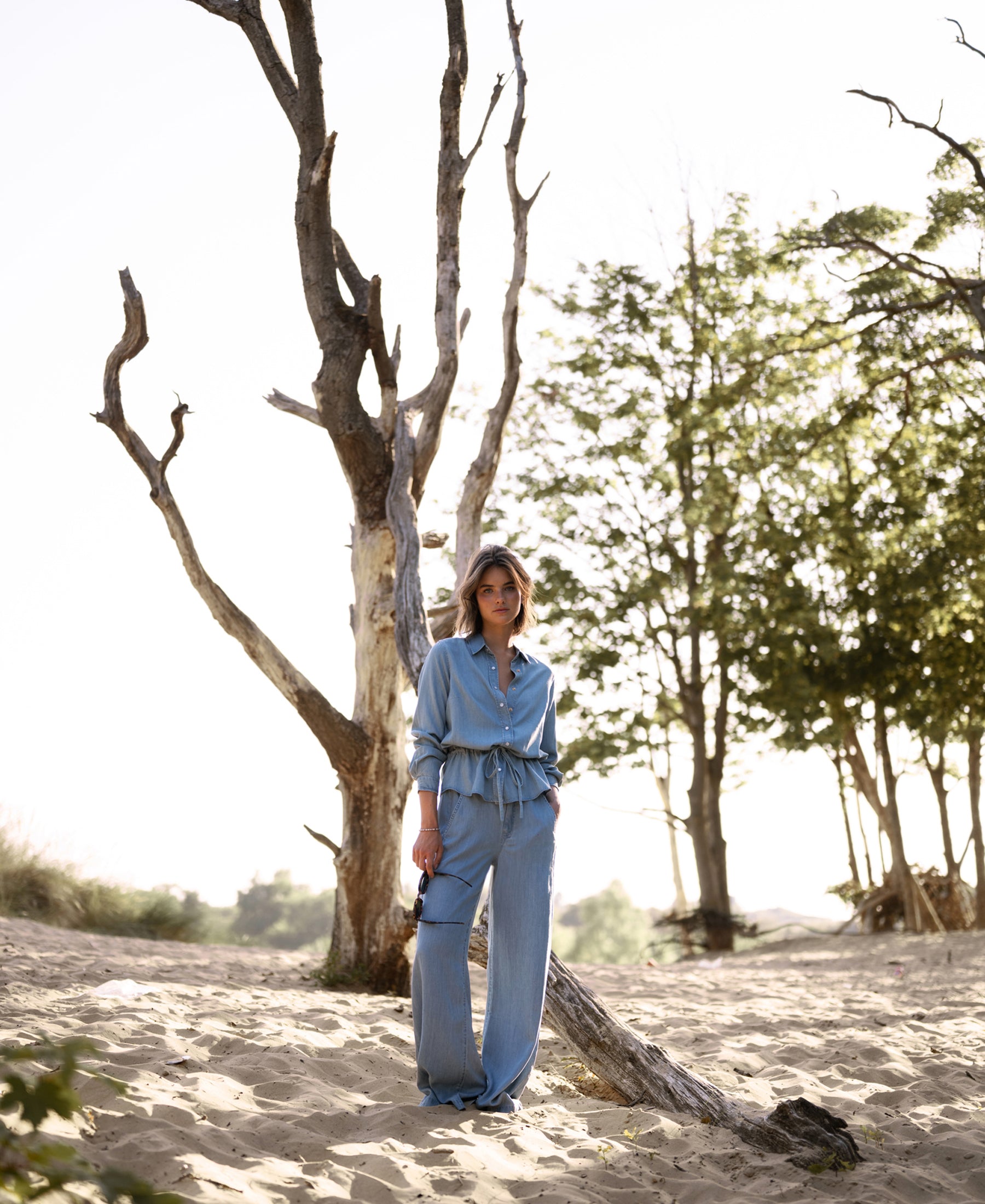 Woman wearing light blue Tencel denim blouse stands outdoors with sand and trees in the background.