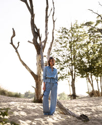 Woman wearing light blue Tencel denim blouse stands outdoors with sand and trees in the background.