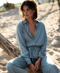 Woman wearing PLEIN PUBLIQUEs Tencel denim blouse sits on sand by a log, looking sideways in sunlight.