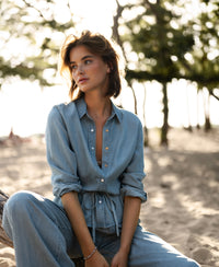 Woman wears light-wash Tencel denim blouse, seated on a sunlit beach log among trees.