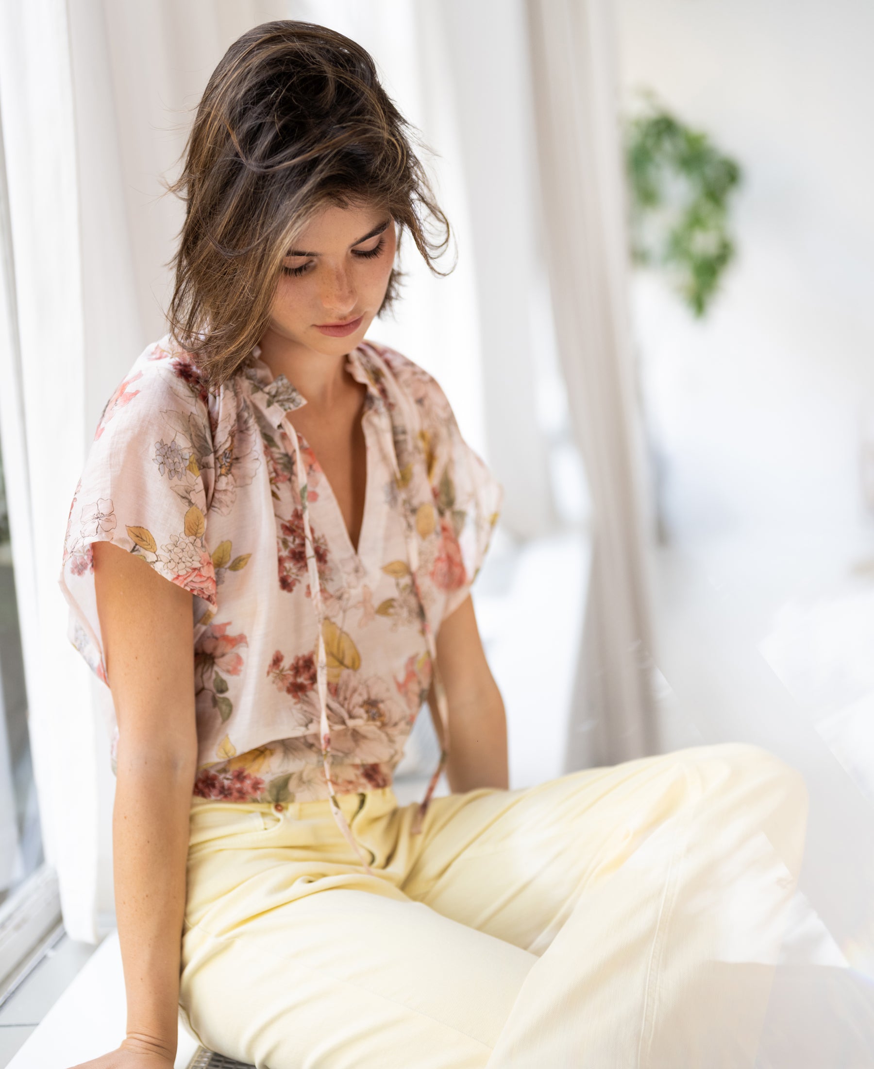 Woman wearing a blush floral print blouse sits calmly by a window.