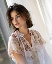 Woman wearing a blush floral print blouse top by PLEIN PUBLIQUE, seen from the side near a window.