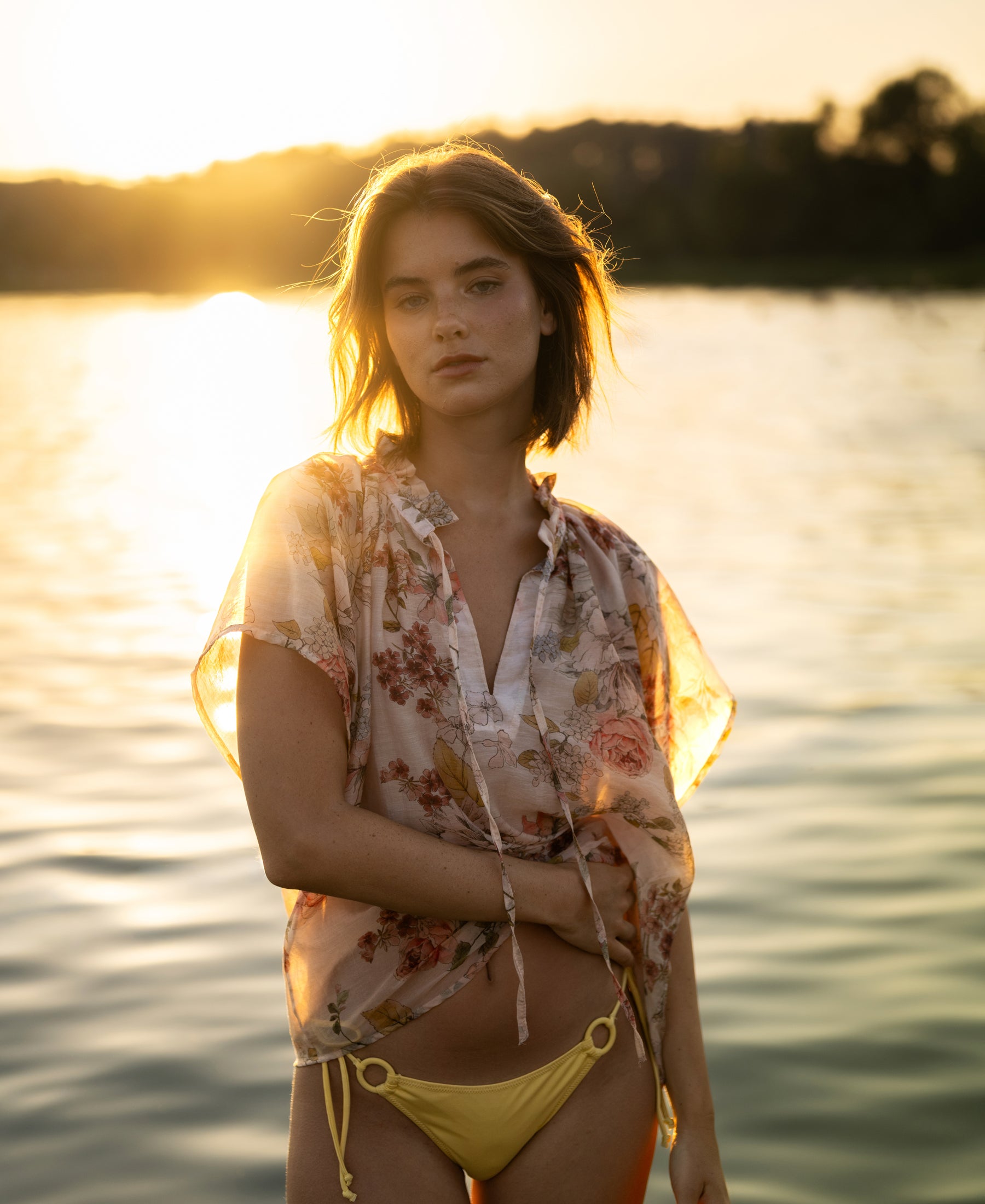 Woman wearing blush floral blouse stands by a lake at sunset, sunlight behind her.