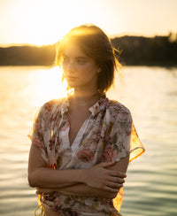 Woman wearing a blush floral print blouse top by PLEIN PUBLIQUE, standing outdoors at sunset.
