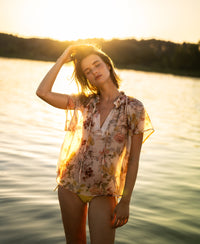 Woman wearing blush floral print blouse stands by water at sunset, eyes closed.