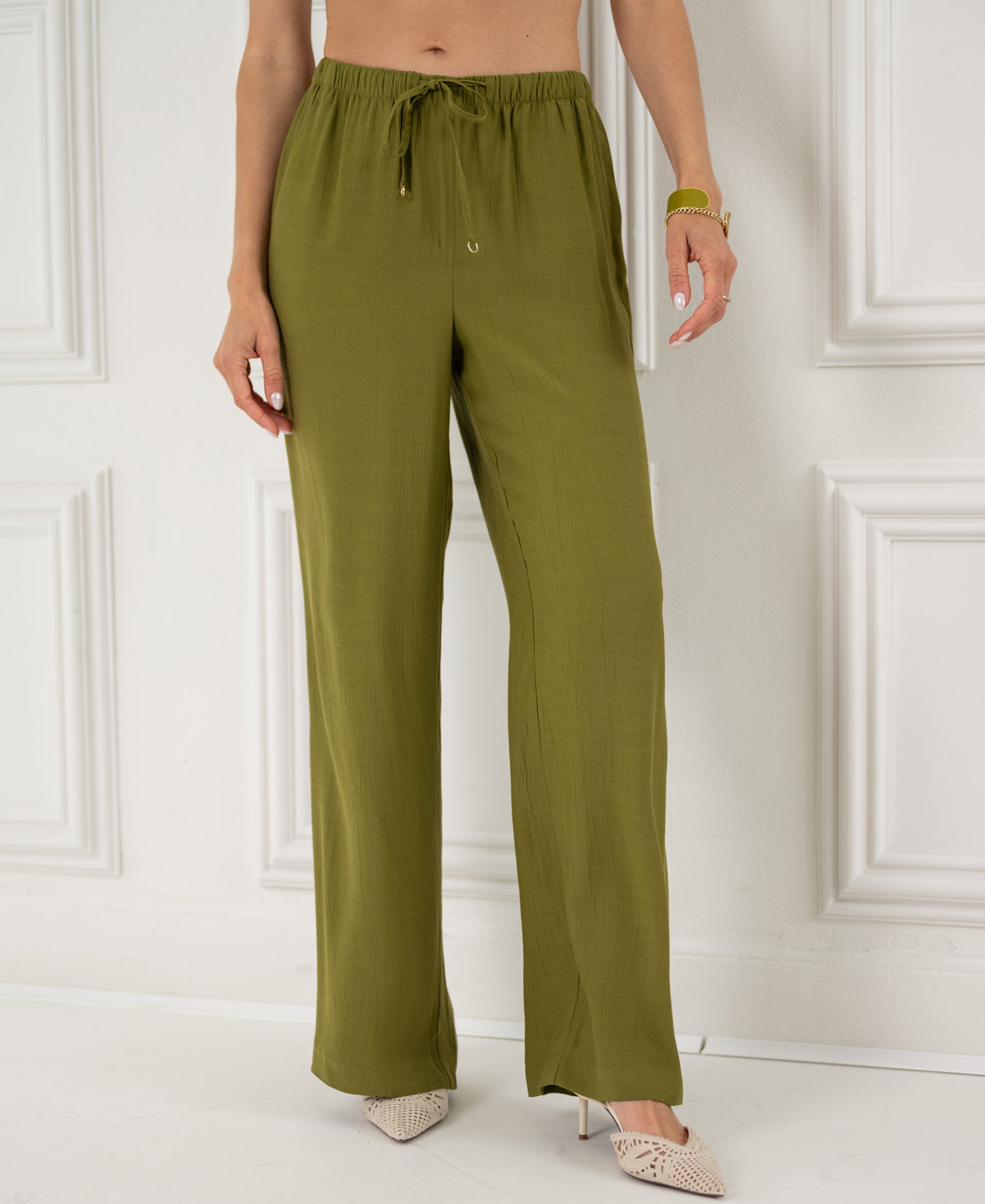 Model wears olive green LA LYNETTE trousers, standing by a white wall.