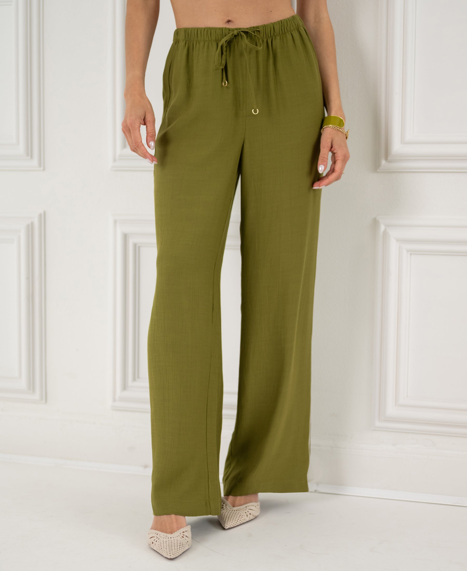 Woman wears olive green LA LYNETTE trousers, pictured standing by a white paneled wall.