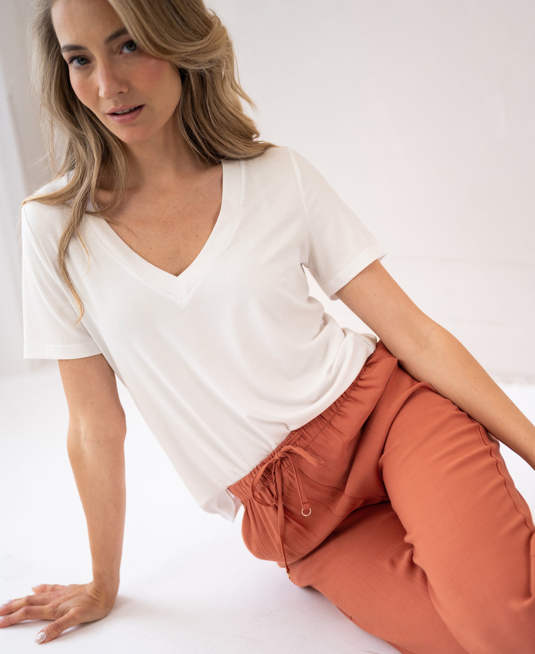 Terra-colored LA LYNETTE pants shown on a model sitting on the floor in a bright, minimal room.