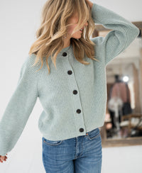 Woman wearing a light green Merino wool vest stands indoors, looking to the side with one hand in her hair.