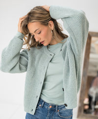 Mist green Merino wool vest by PLEIN PUBLIQUE, shown indoors. Model looks down with hands in her hair.
