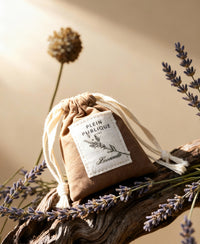 A small brown lavender sachet by PLEIN PUBLIQUE lies on wood with lavender.