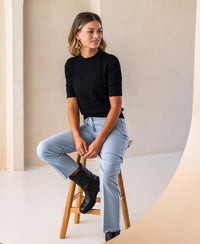 Black merino wool sweater, shown on a woman seated in a minimal studio.