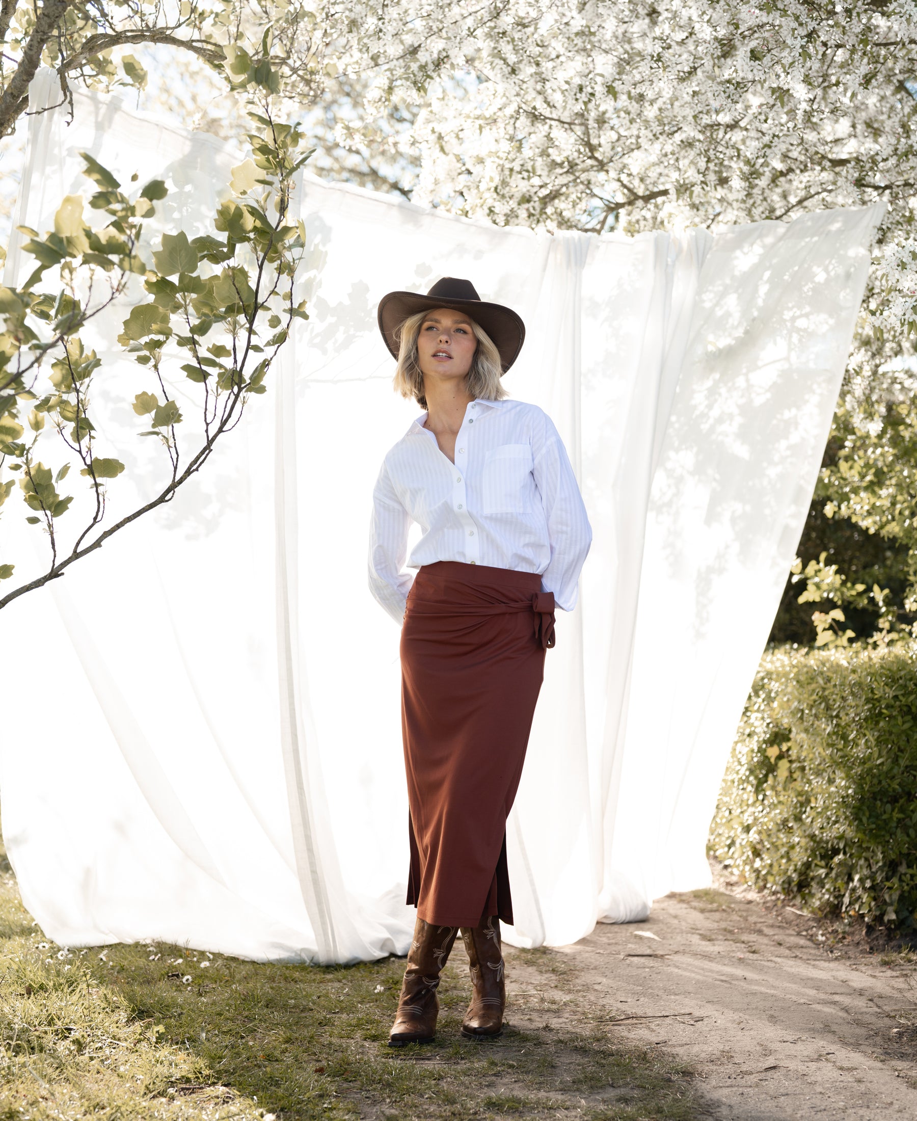 Camel Travel skirt LE MAEL by PLEIN PUBLIQUE shown near a white curtain and tree.