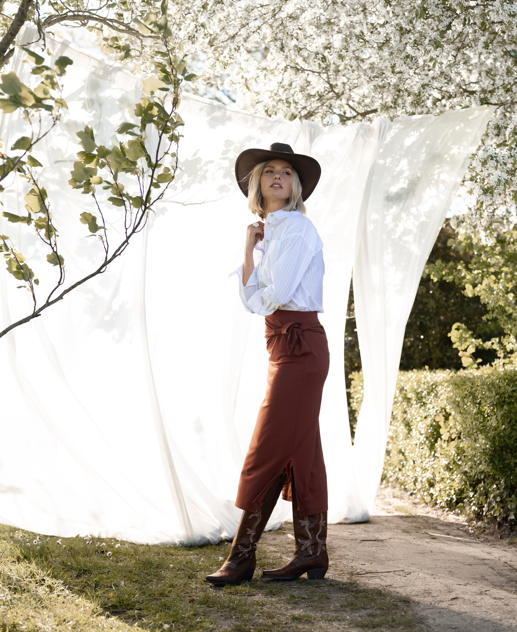 Camel Travel Skirt LE MAEL by PLEIN PUBLIQUE, shown outdoors by white drapes and trees.