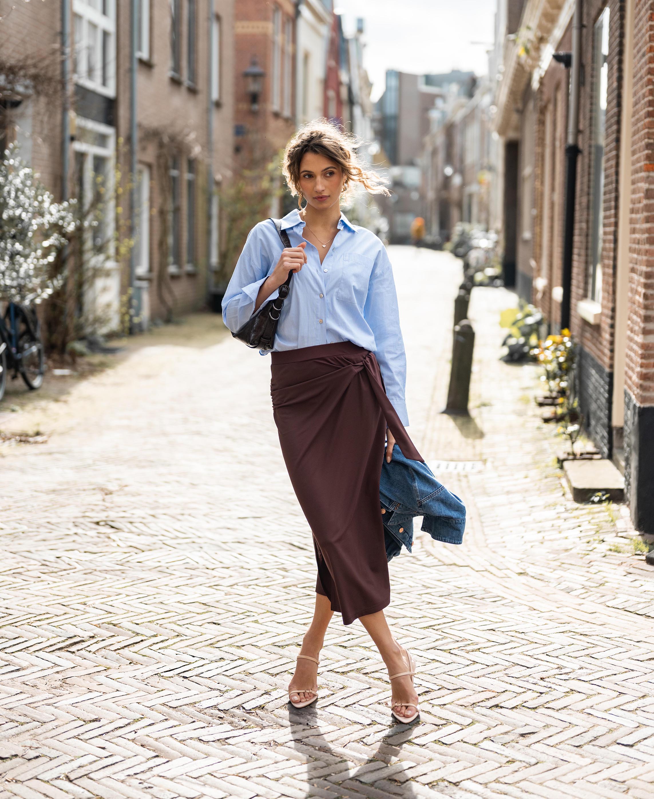 Woman wearing a brown LE MAEL Travel skirt by PLEIN PUBLIQUE, standing on a cobblestone street.