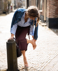 A woman wears the Travel Skirt LE MAEL in cacao brown by PLEIN PUBLIQUE, shown outdoors on a sunny street.