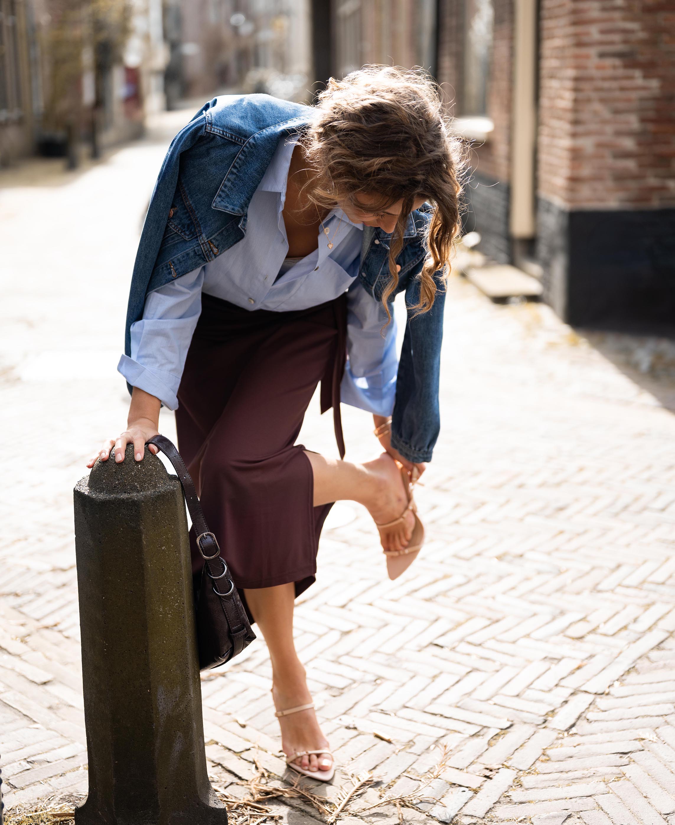 A woman wears the Travel Skirt LE MAEL in cacao brown by PLEIN PUBLIQUE, shown outdoors on a sunny street.
