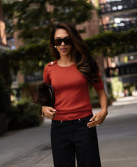 Woman wearing a Merino wool sweater in chili red, outdoors with greenery and buildings.