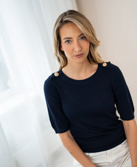 Navy blue merino wool sweater by PLEIN PUBLIQUE, shown on a model standing near a window with white curtains.