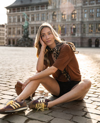 Woman wearing the PLEIN PUBLIQUE silk top LE MARDY Havana sits on a cobblestone square.