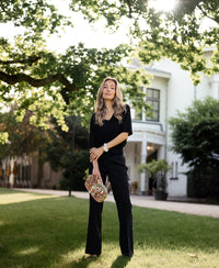 Woman wearing a black silk top by PLEIN PUBLIQUE stands on grass near a white house.