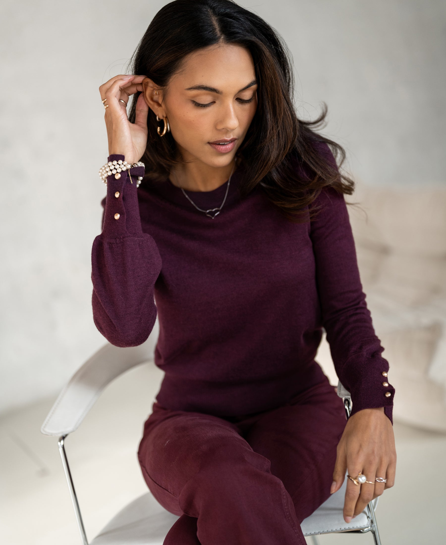 Woman wearing PLEIN PUBLIQUE merino wool sweater LA MARIS in wine-gold sits on a chair, touching her hair.