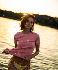 Woman wearing a soft Luxe T-shirt in light flamingo pink by Plein Publique stands in sunset water.