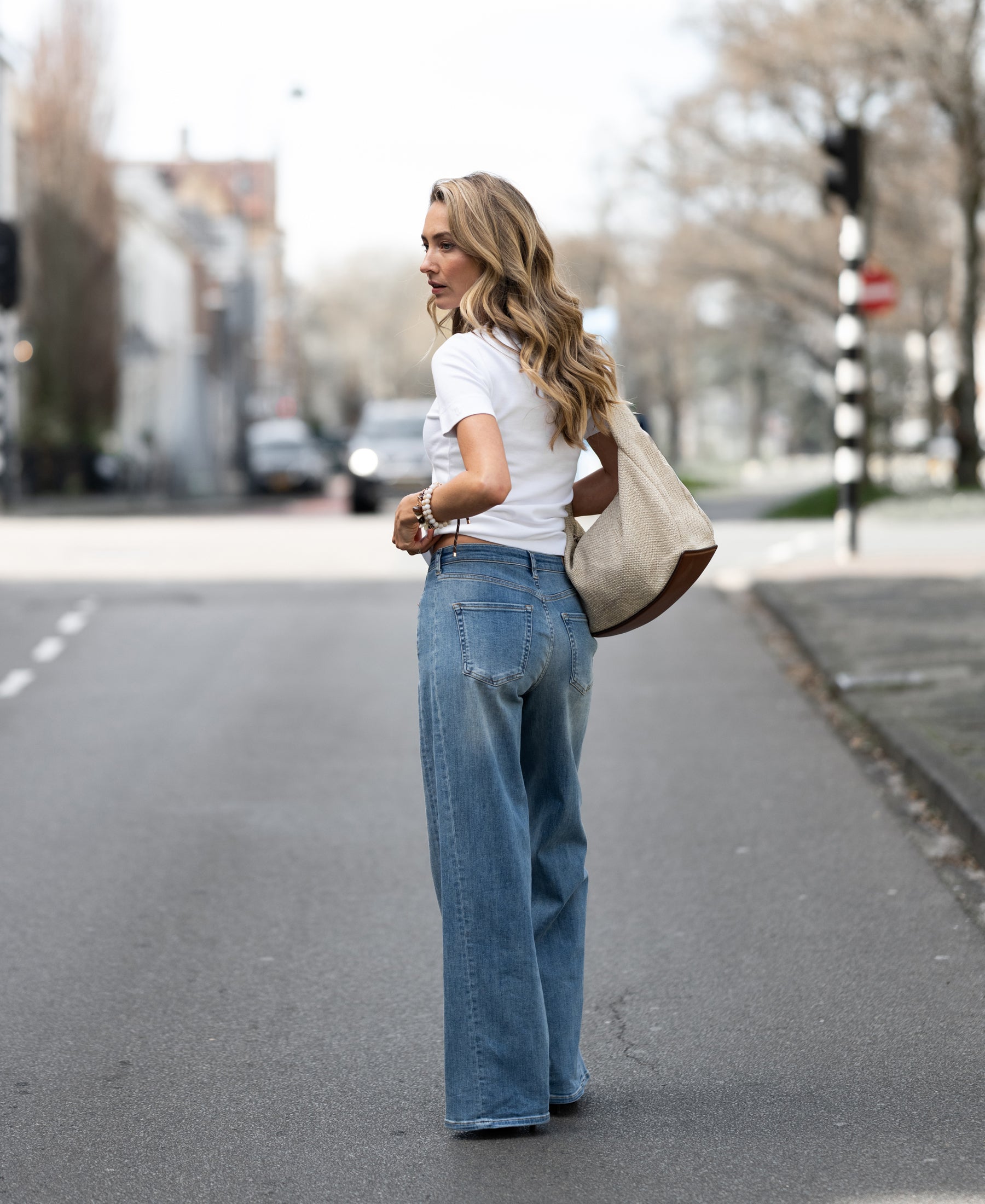 Woman wearing wide leg light stone wash jeans stands on the street, looking back.