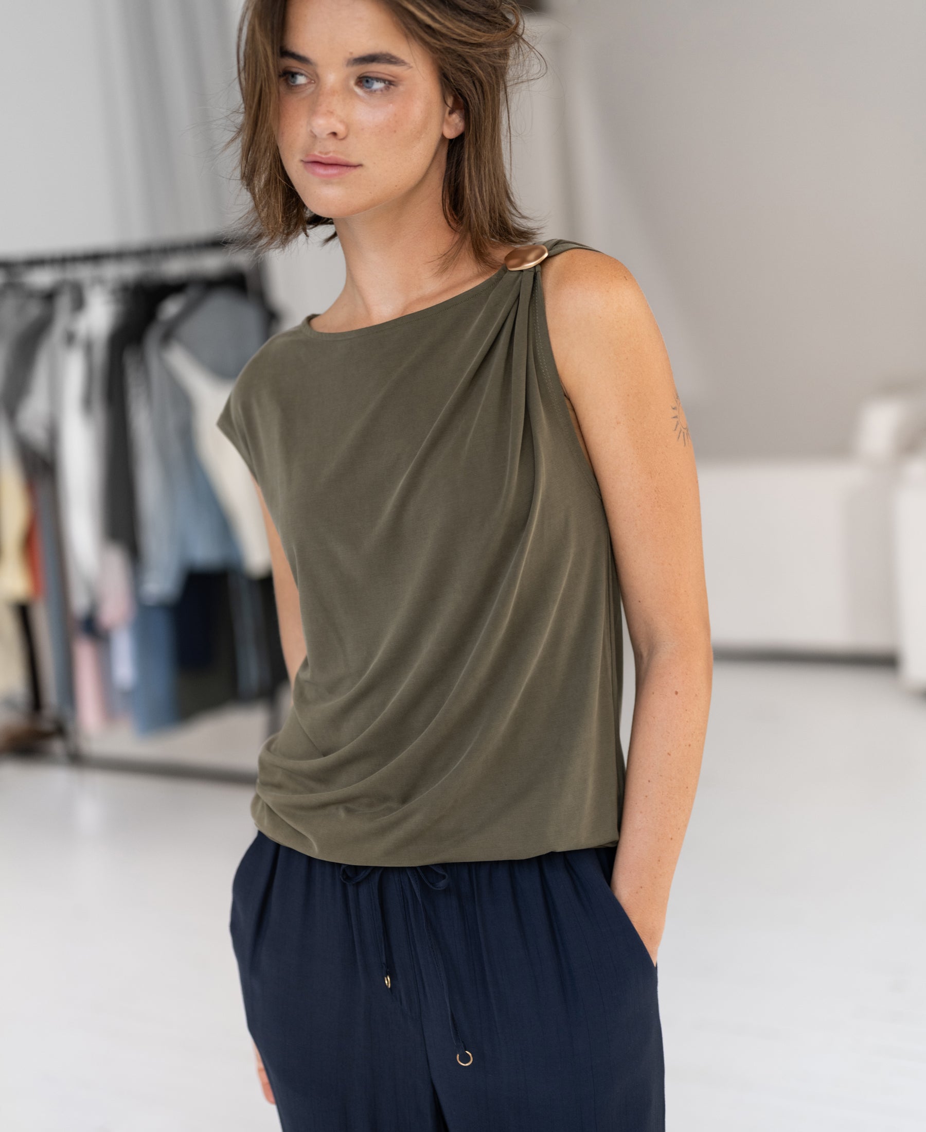 A woman indoors wears the LA MAYA Olive Green Top with buckle by PLEIN PUBLIQUE; clothes hang behind her.
