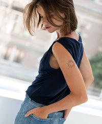 Woman wearing dark blue PLEIN PUBLIQUE LA MAYA top with buckle, sun tattoo on upper arm, hands in pockets.
