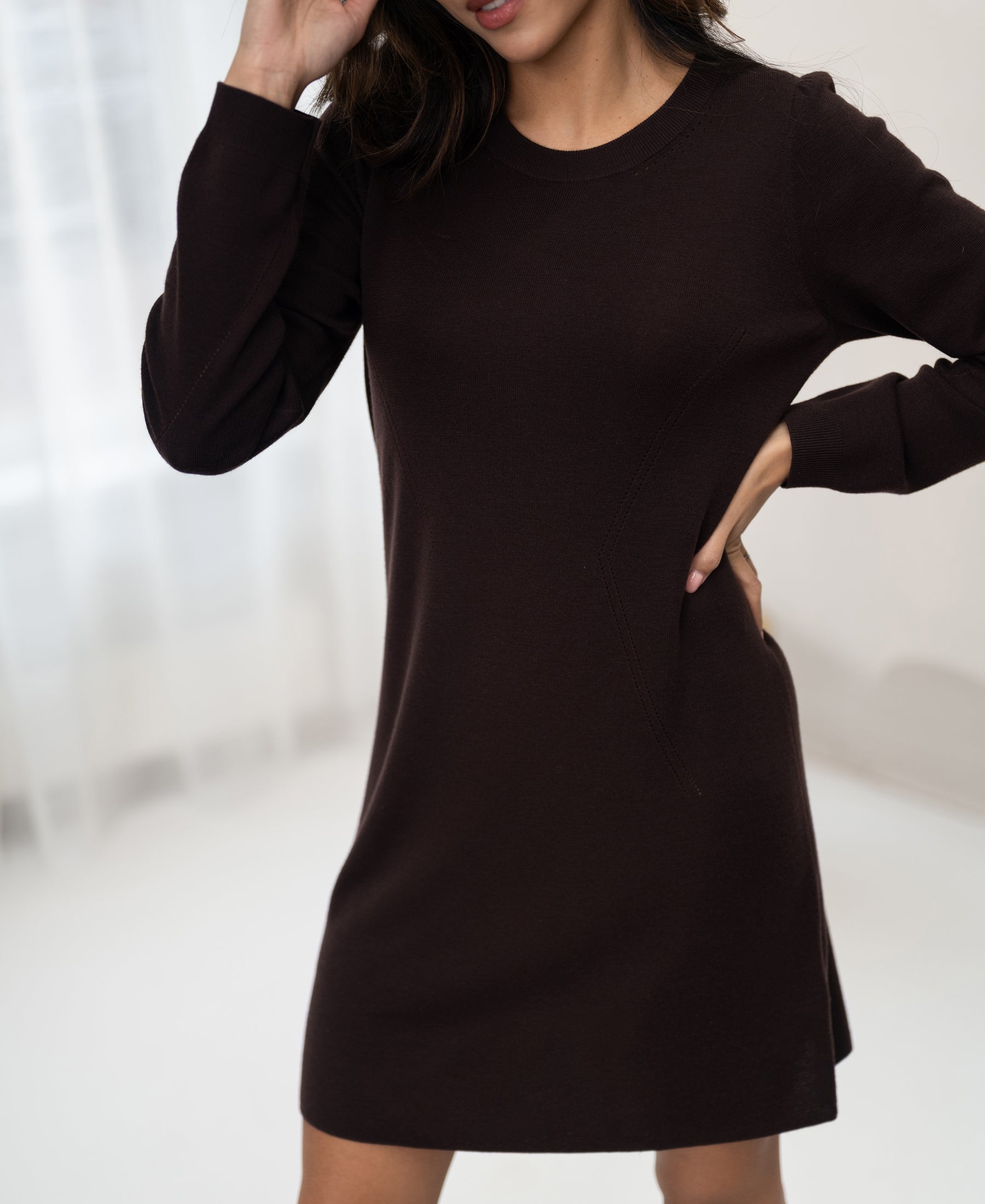 Woman wearing the PLEIN PUBLIQUE brown merino wool dress LA MIMI stands indoors, hand on hip and near her head.