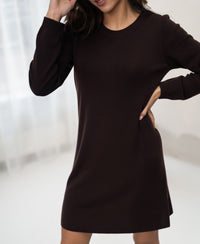 Woman wearing the PLEIN PUBLIQUE brown merino wool dress LA MIMI stands indoors, hand on hip and near her head.