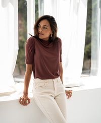 Woman wearing ribbed collared top in cocoa brown stands by a sunlit window, looking to the side.