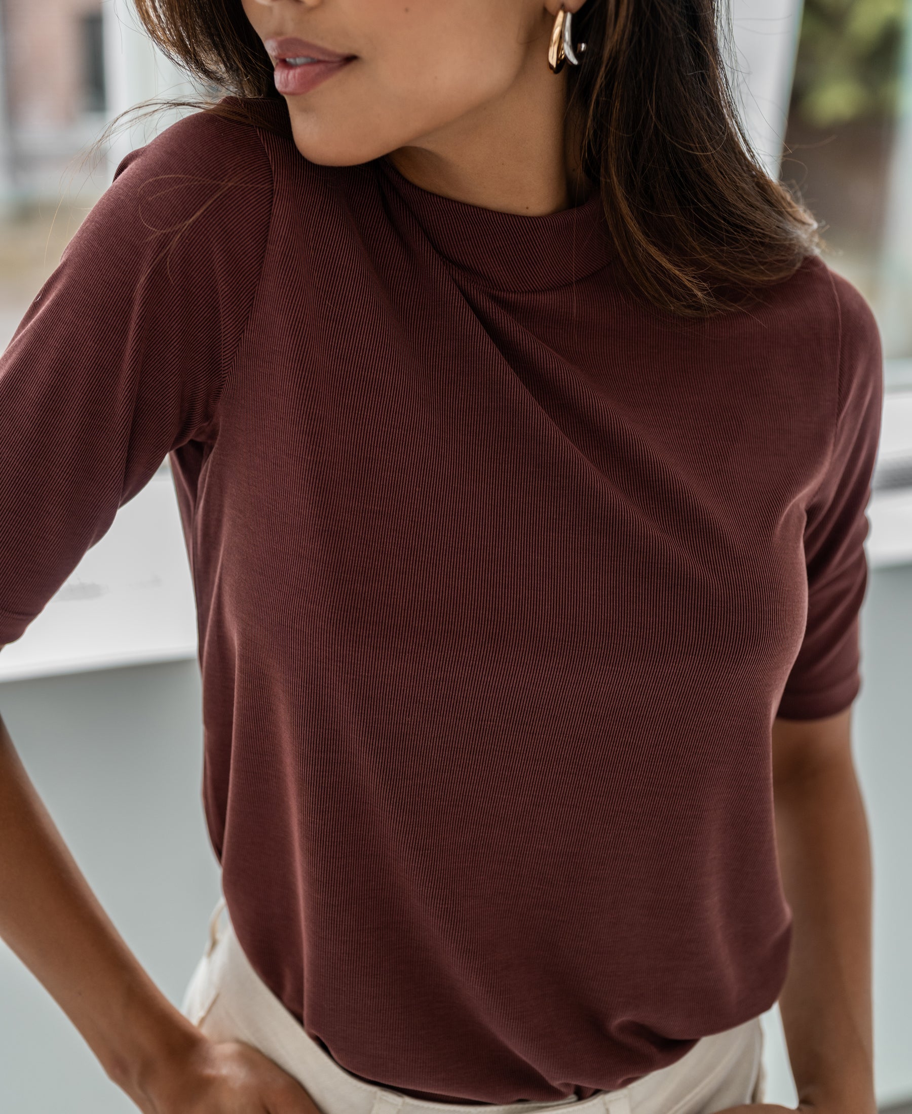Woman wearing a ribbed collared top in cacao color, by Plein Publique, standing indoors near a window.